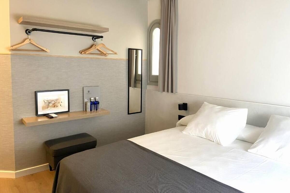 Ein modernes Hotelzimmer mit einem Bett, einem Nachttisch, einem Spiegel, einem an der Wand befestigten Regal mit Bilderrahmen und Telefon sowie einem Kleiderhaken.