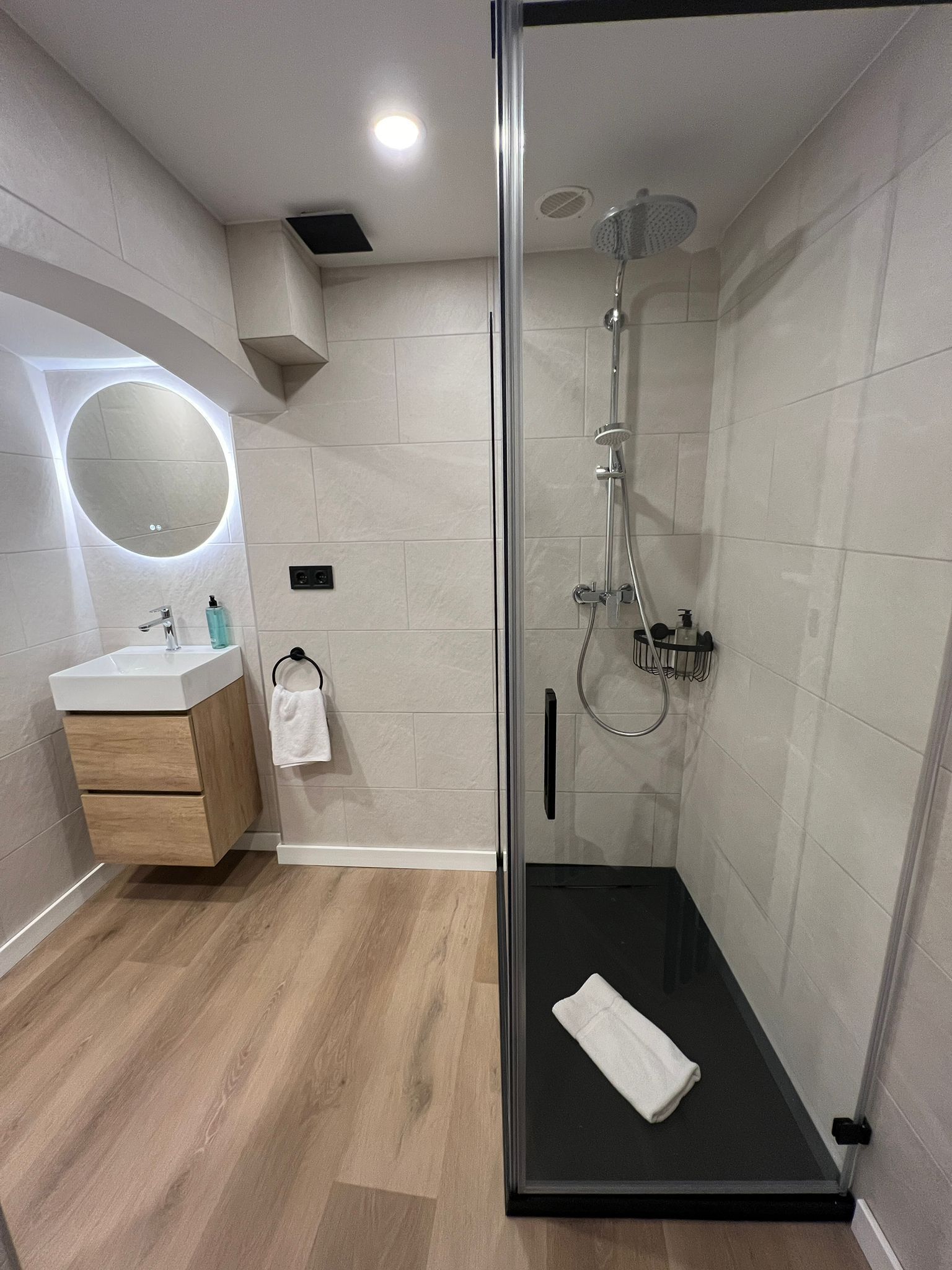 Een moderne badkamer met een douchecabine van glas, een ronde spiegel, een houten wastafelmeubel en een witte handdoek.