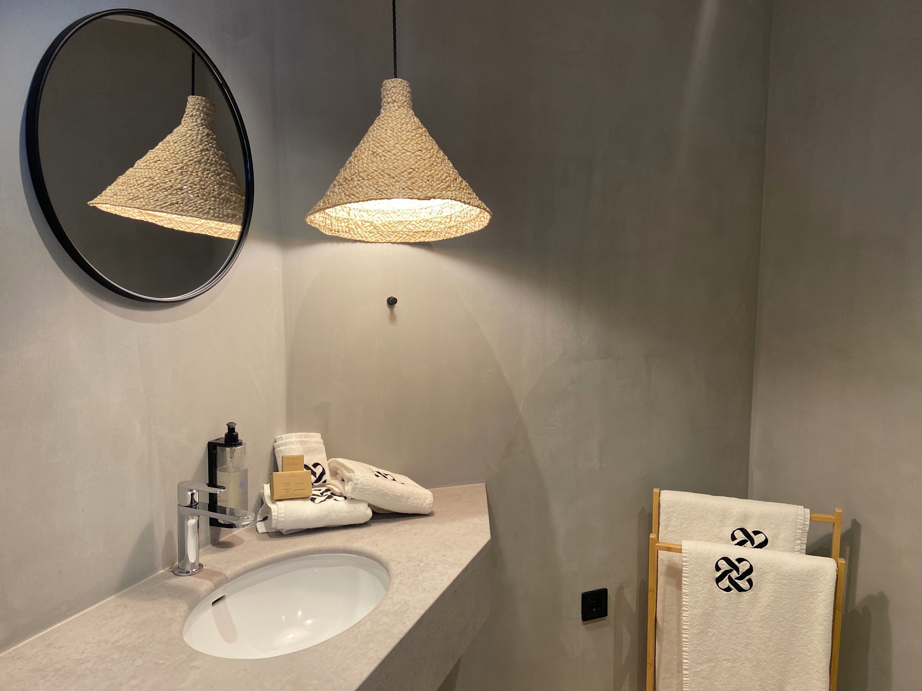 A bathroom with a white sink, a round mirror, two woven pendant lights, and a towel rack with towels.