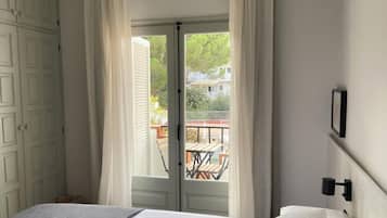 Un dormitorio con cama, una ventana con cortinas y vistas a un balcón con mobiliario exterior, además de una lámpara fijada a la pared.