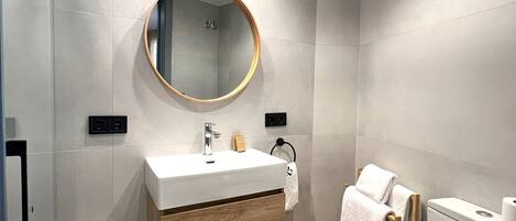 A modern bathroom with a round mirror, white sink, wooden vanity, and a toilet.