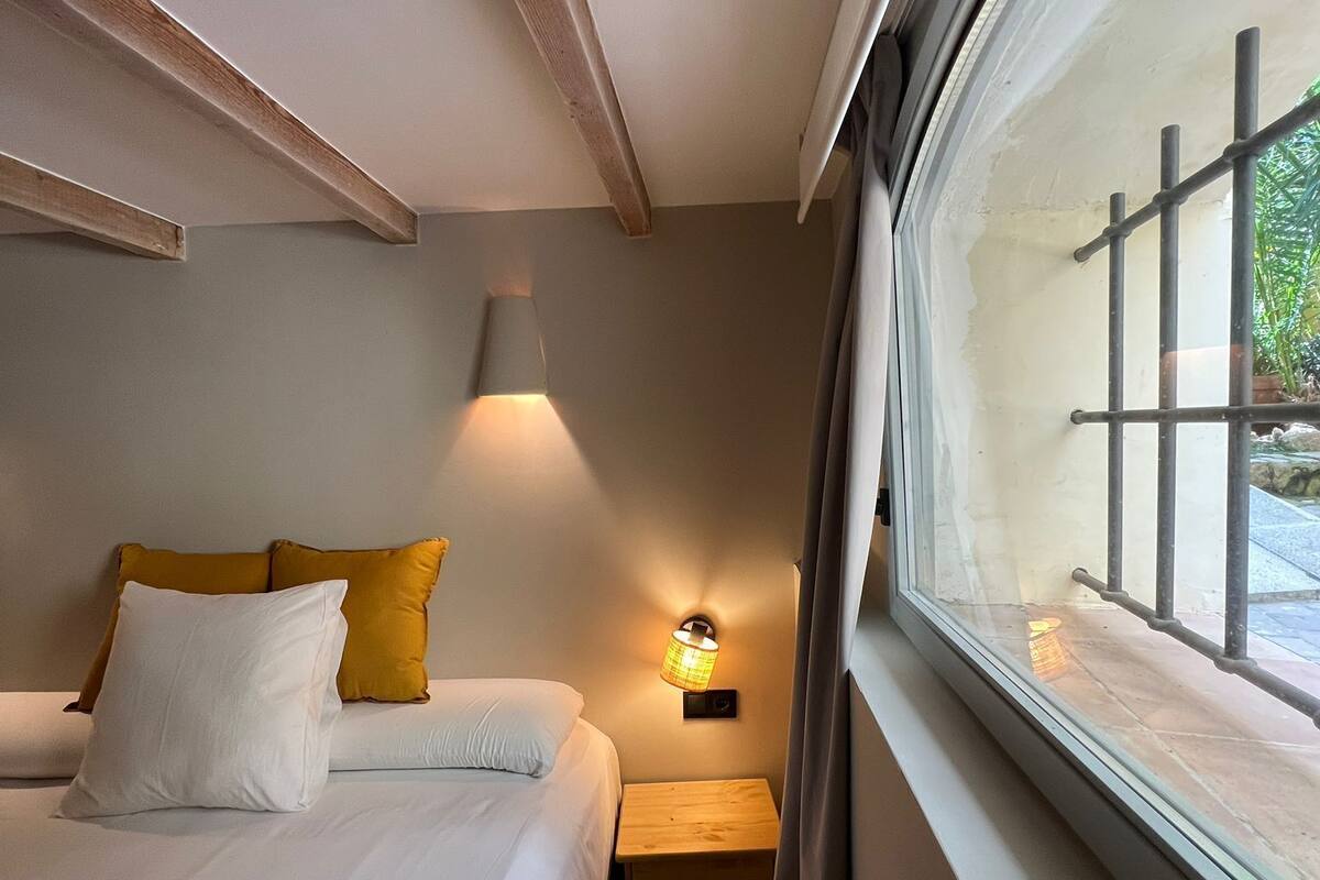 Ein kleines, gemütliches Schlafzimmer mit Bett, Nachttischlampe und Fenster mit Blick ins Freie.