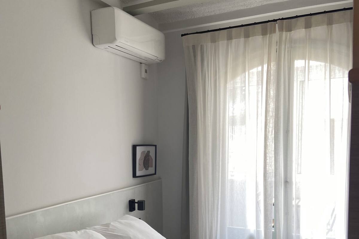 Ein ordentlich bezogenes Bett mit weißen Laken, einer grauen Tagesdecke und einem Kopfteil mit Wandleuchten.