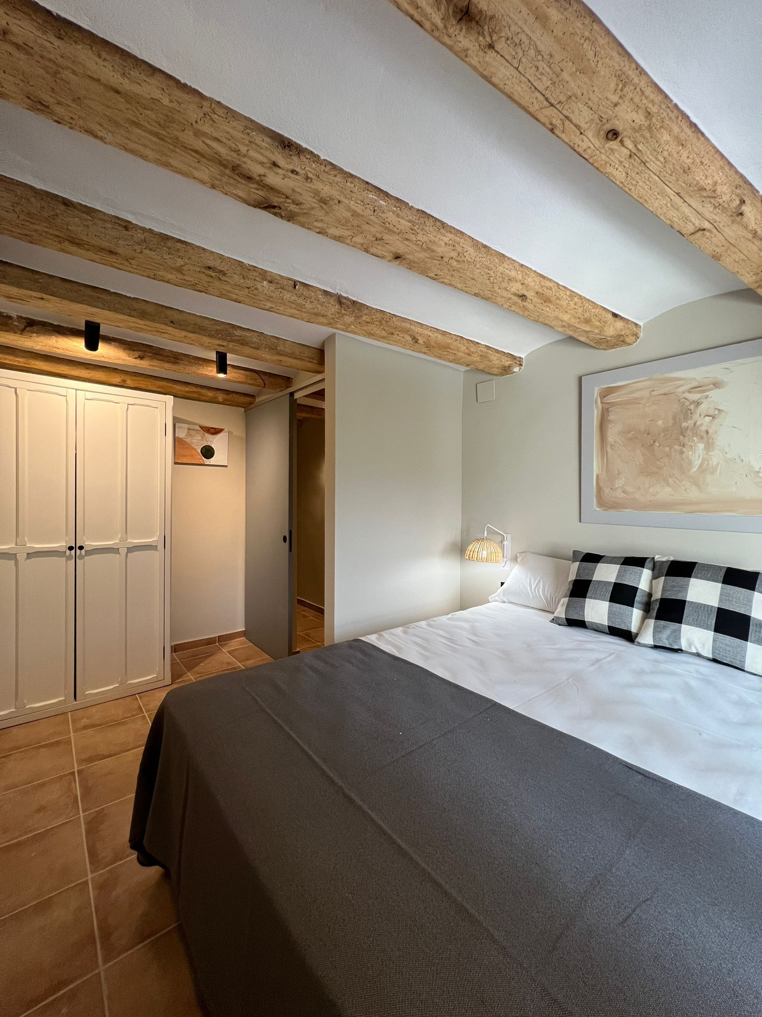 A bedroom with a bed, checkered pillows, a wall-mounted lamp, and a painting.