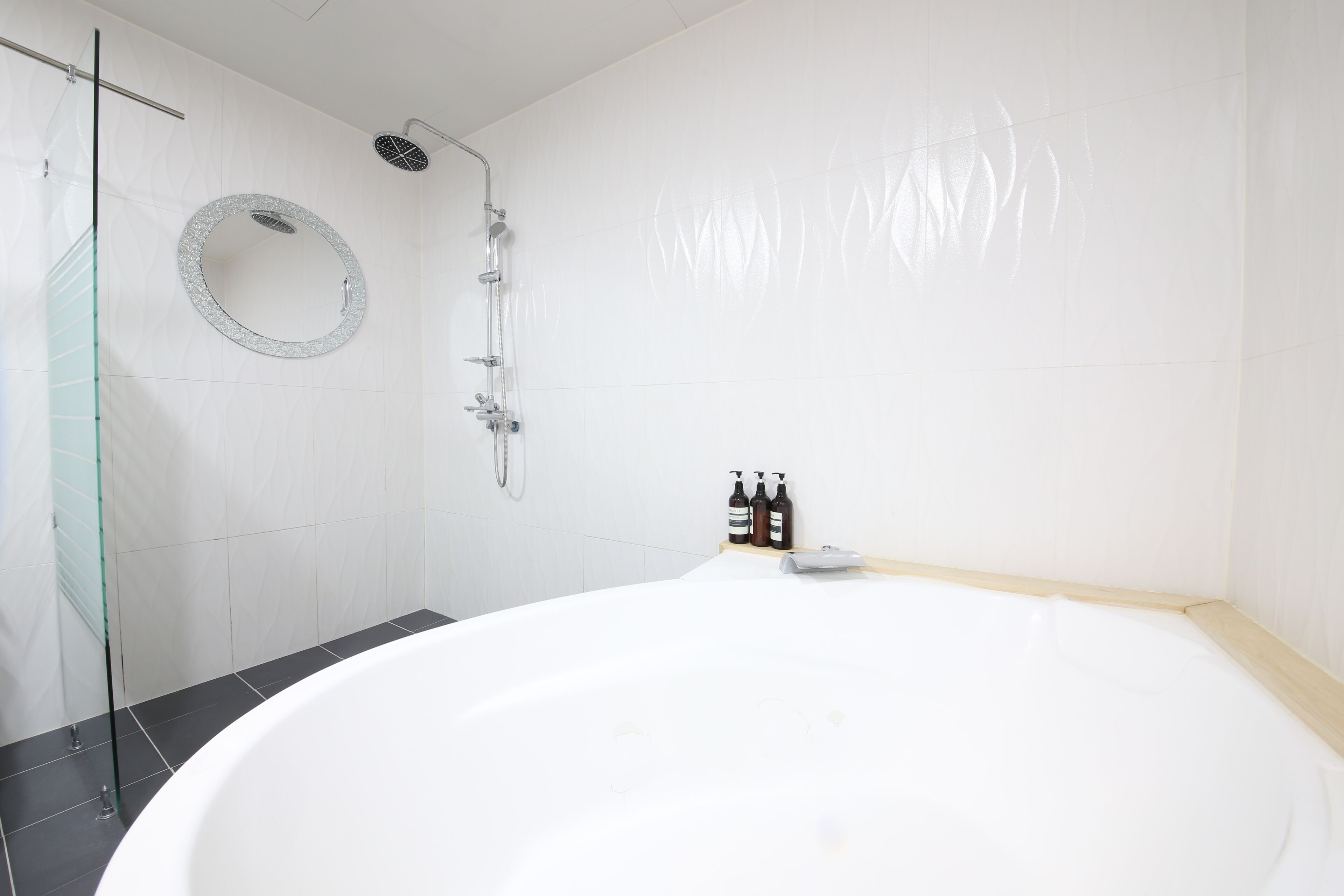 A modern bathroom with a large white bathtub, a glass shower enclosure, and a round mirror on the wall.