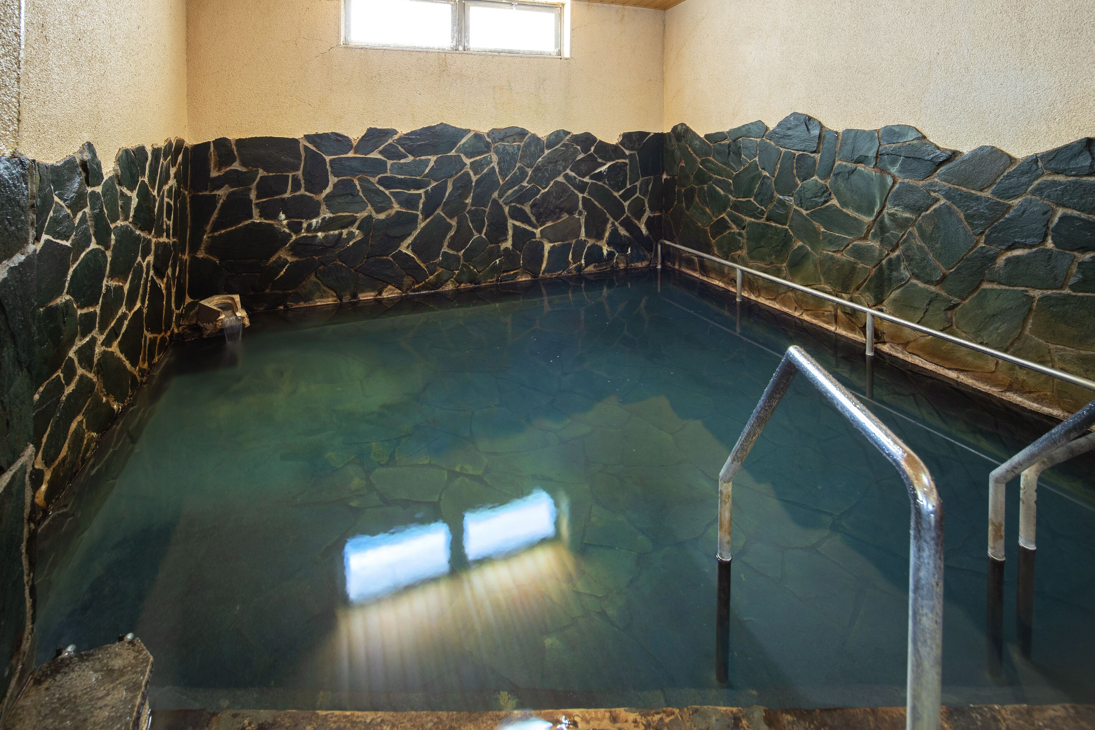 Una piscina techada con paredes de piedra, agua cristalina, barandillas de metal y un ventanal.