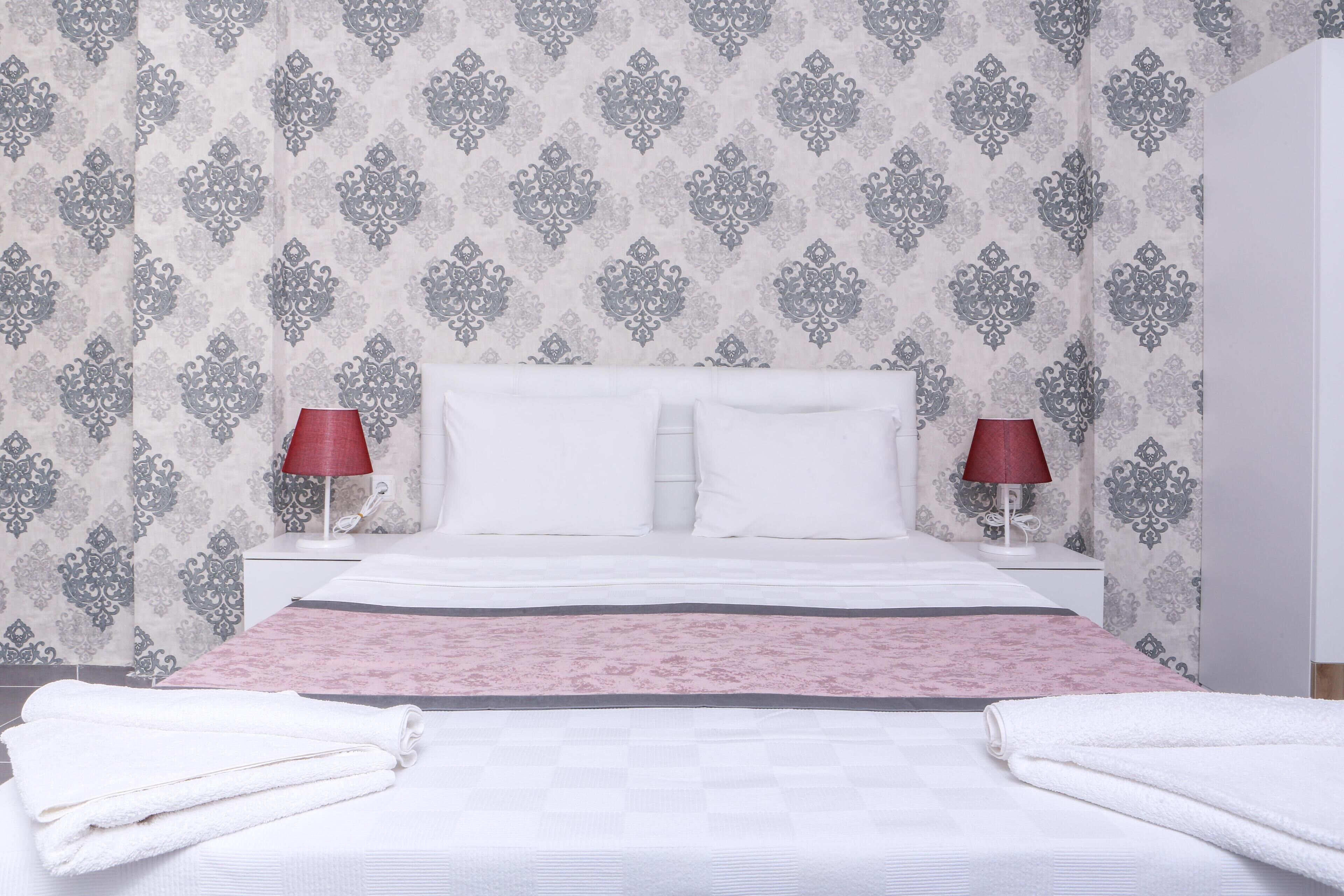 A neatly made bed with white linens and a pink accent, two bedside lamps, and a patterned wallpaper.