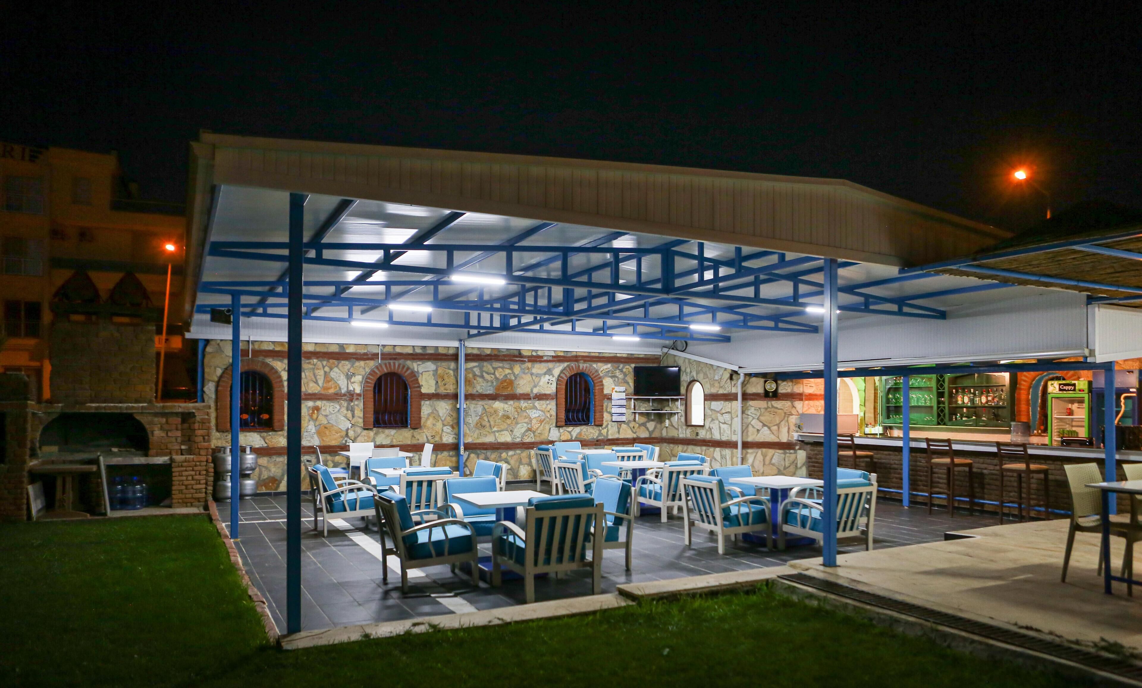 Un patio al aire libre con mesas y sillas, un muro de piedra con ventanas arqueadas y una zona de bar con un letrero verde.
