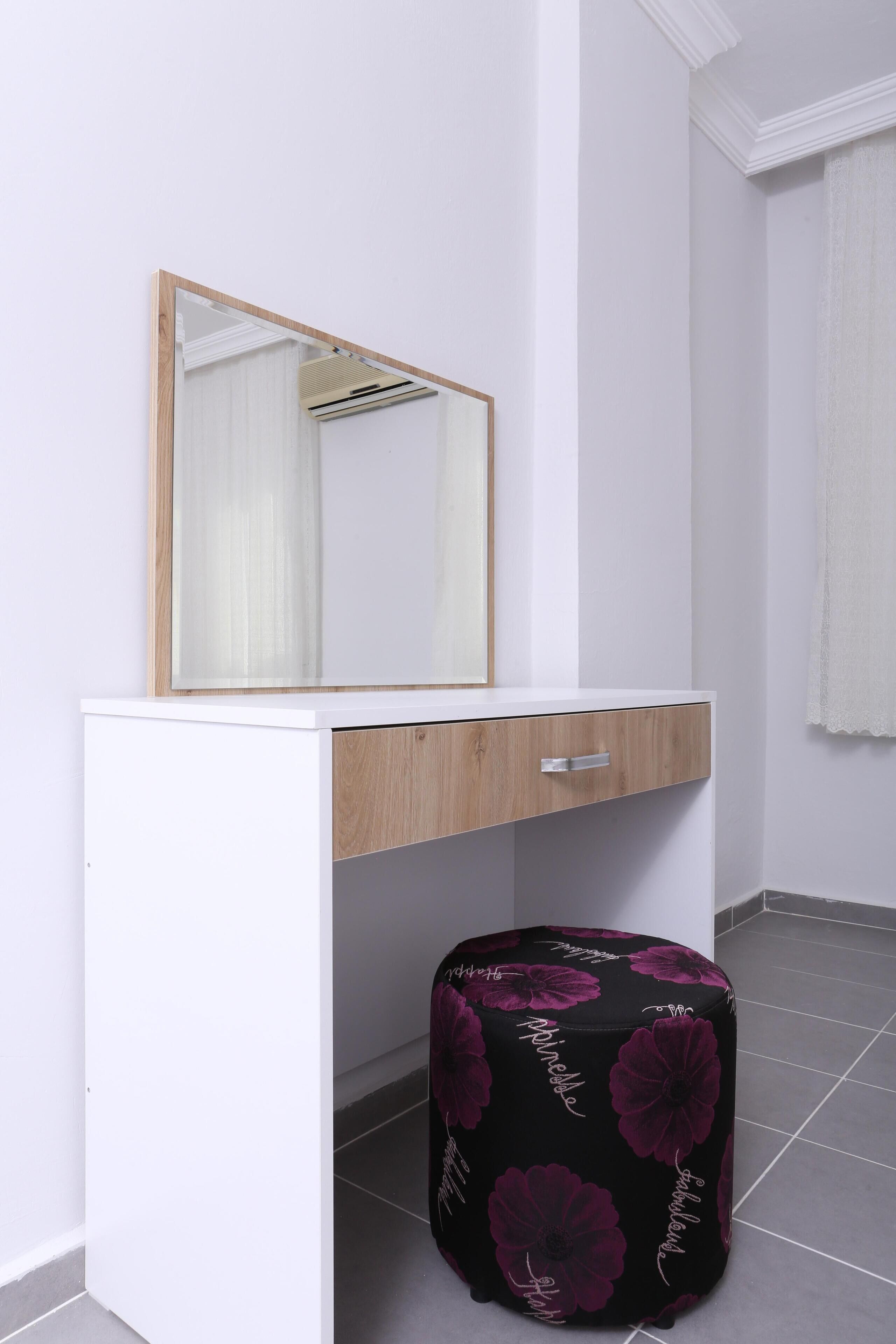 A white vanity with a mirror and a wooden drawer, a black ottoman with floral patterns, and a tiled floor.
