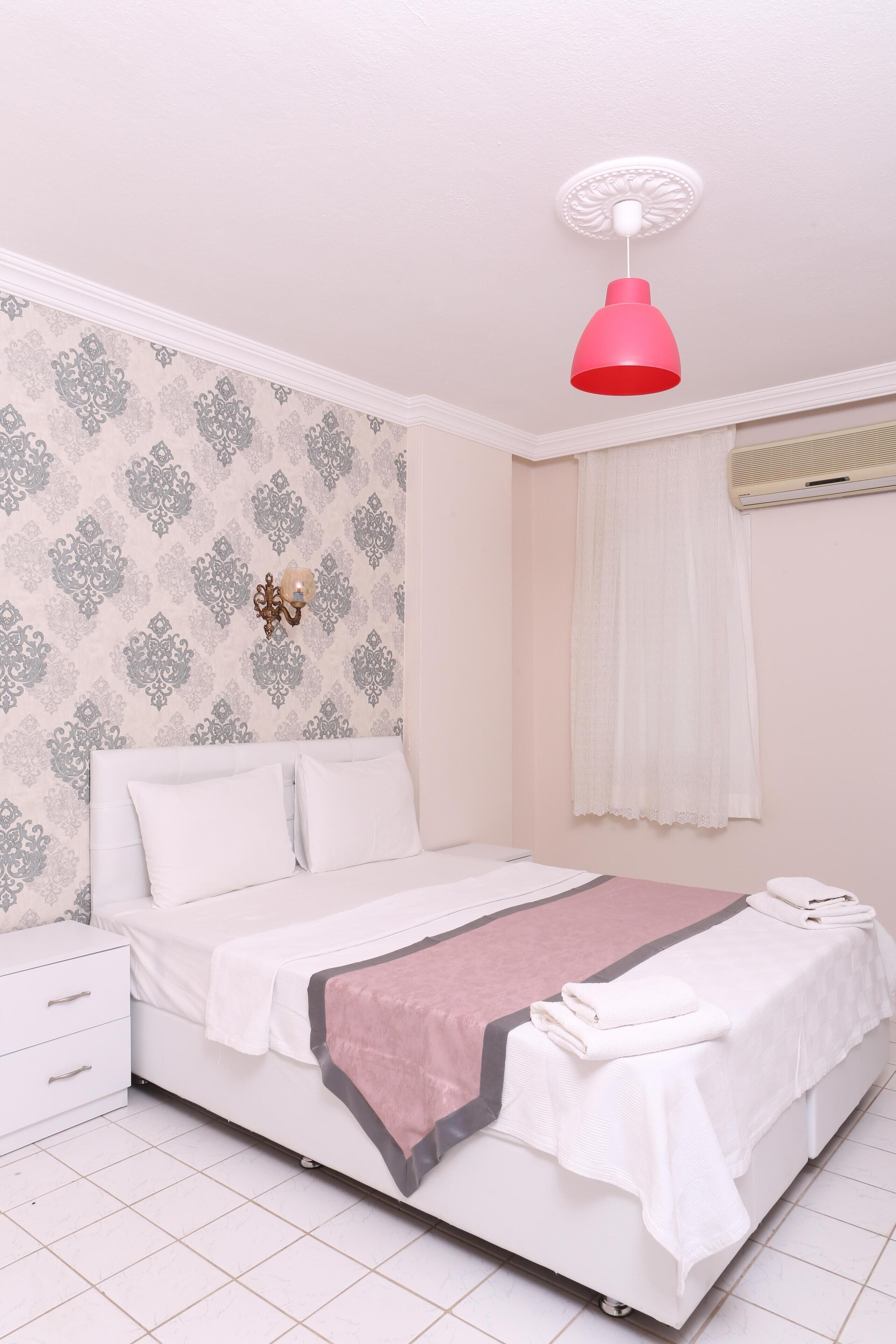 Una cama bien hecha con una manta blanca y rosa, una mesita de noche y una lámpara de pared.