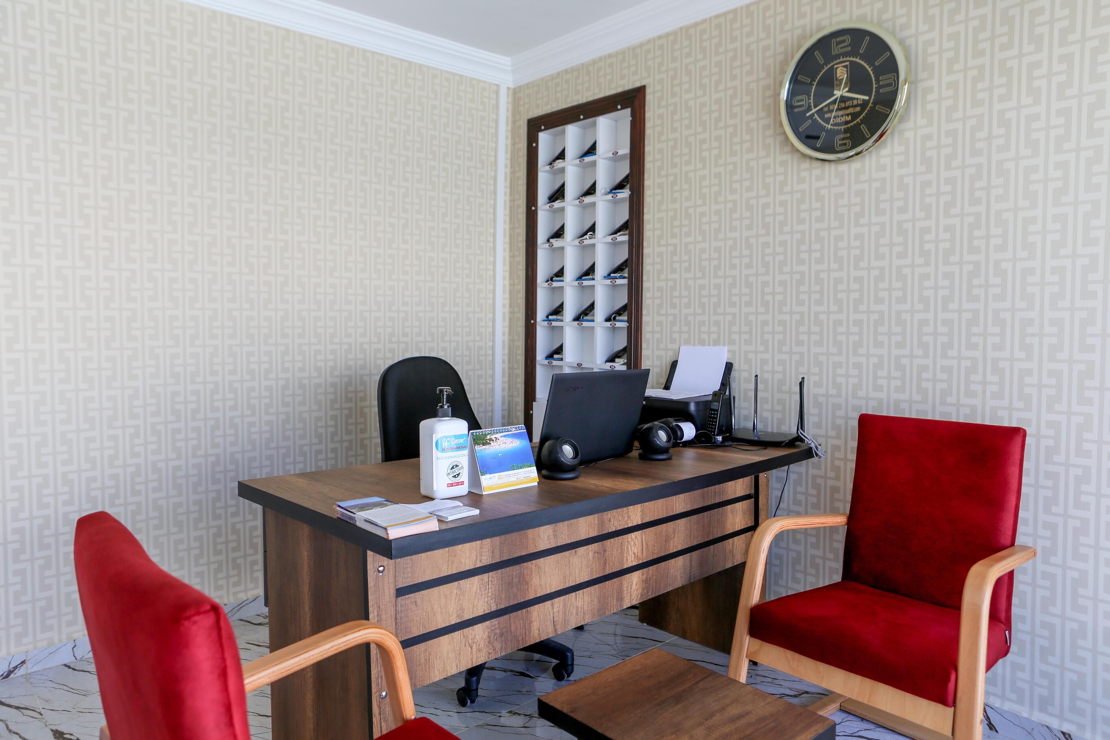 A hotel room with a wooden desk, two red chairs, a computer, and a wall clock.