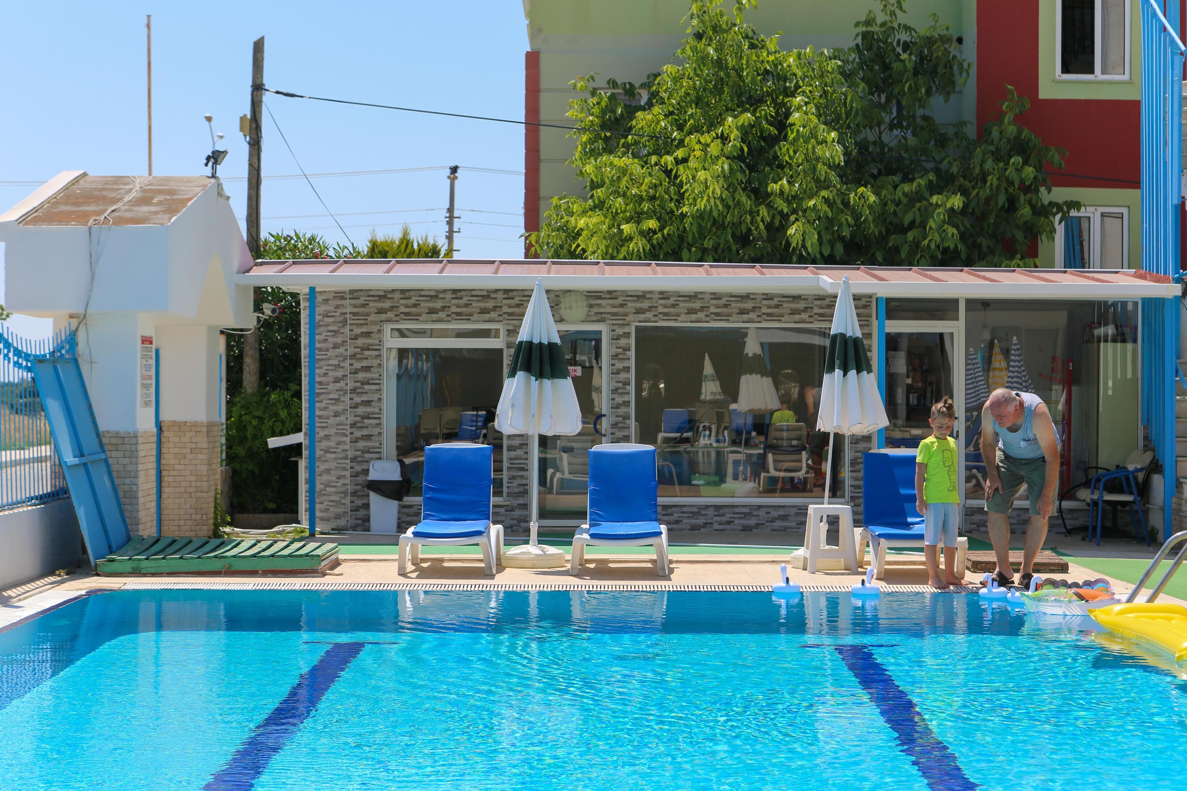 A swimming pool with lounge chairs and umbrellas, a small building with windows, and a tree in the background.