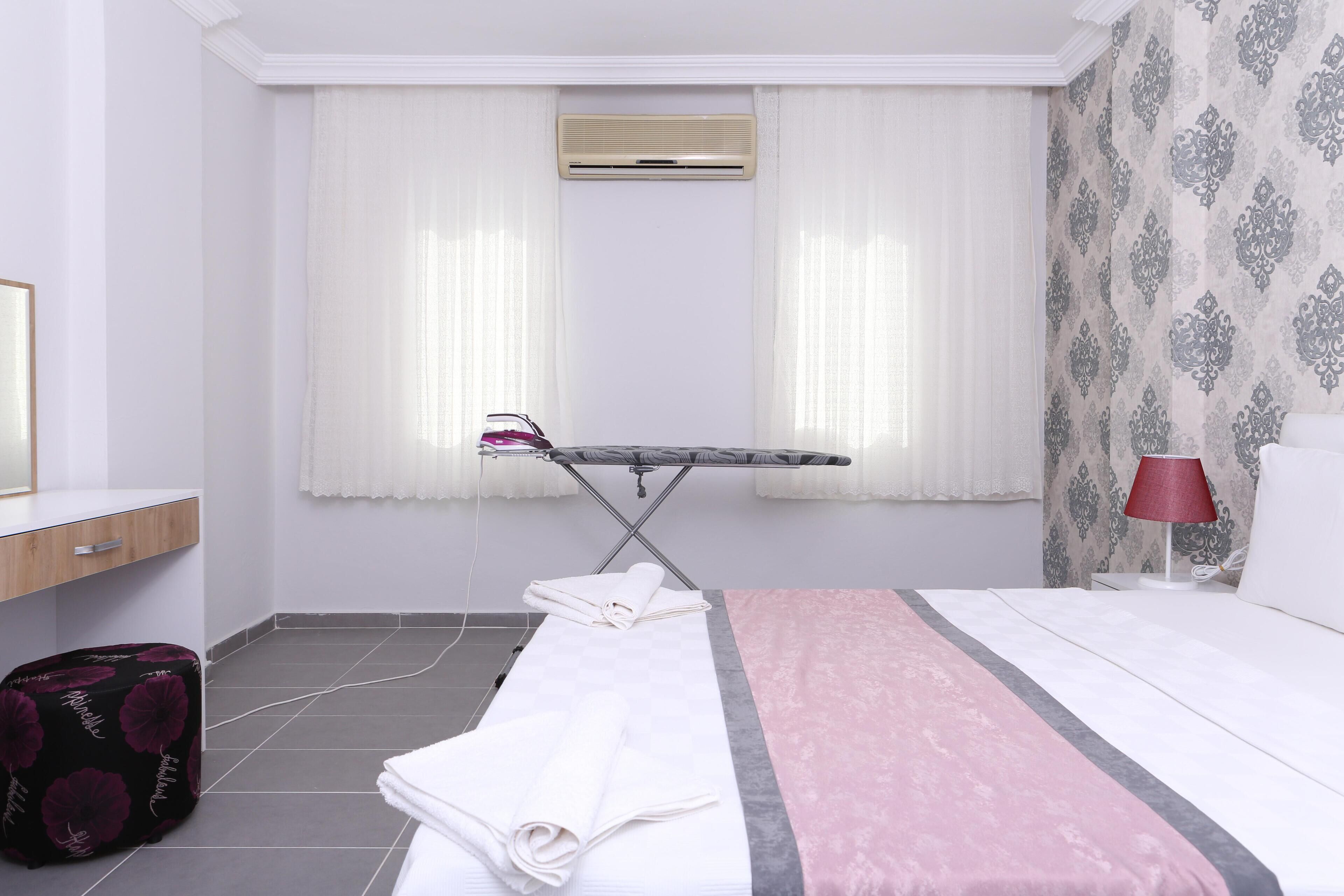 A hotel room with a bed, an ironing board, a suitcase, and a window with curtains.