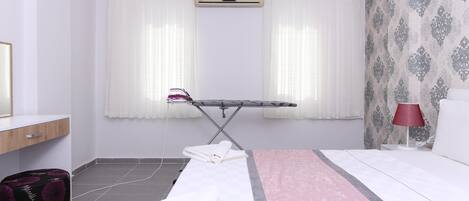 A hotel room with a bed, an ironing board, a suitcase, and a window with curtains.