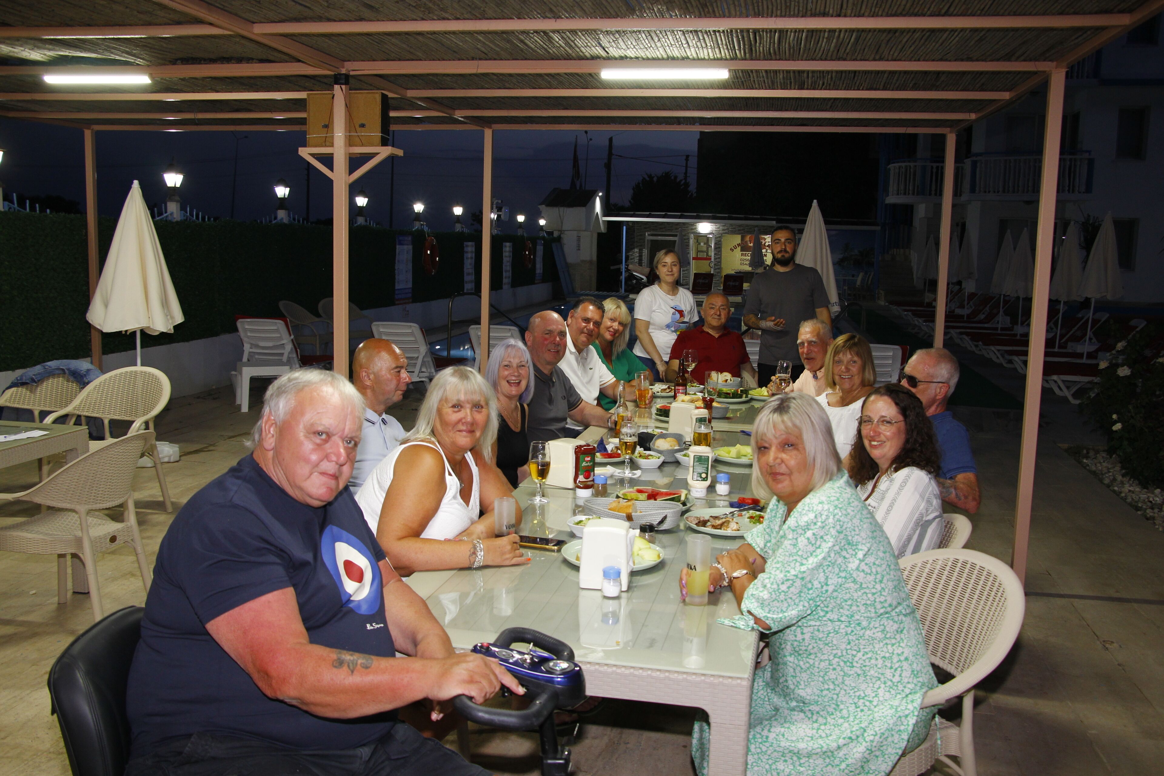A group of people seated around a table in an outdoor covered area with chairs and umbrellas.