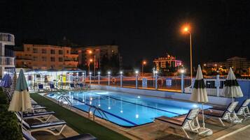 A swimming pool with lounge chairs and umbrellas, surrounded by a fence and illuminated by street lamps.