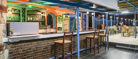 Zona de bar con pared de ladrillo, estanterías verdes con botellas y un refrigerador de cerveza Cappy. Hay taburetes de bar y una mesa con sillas.