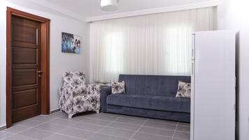 Un salon avec un canapé bleu foncé, un réfrigérateur blanc, une porte en bois et un fauteuil à motifs.