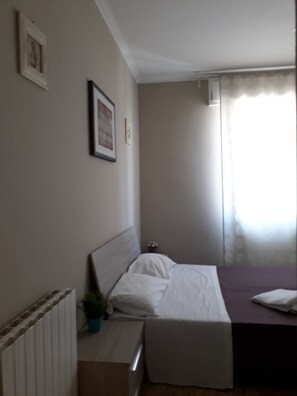 A bedroom with a bed, a bedside table with a plant, a radiator, and a window with curtains. - 48 airport budget rooms (Pisa)