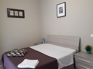 A neatly made bed with a checkered pillow, a white blanket, and a small bedside table with a lamp. There are framed pictures on the wall above the bed. - 48 airport budget rooms (Pisa)