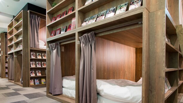 A hotel room with bunk beds, wooden headboards, and shelves with magazines. - Comics & CapsuleHotel COMICAP Sapporo (Sapporo)