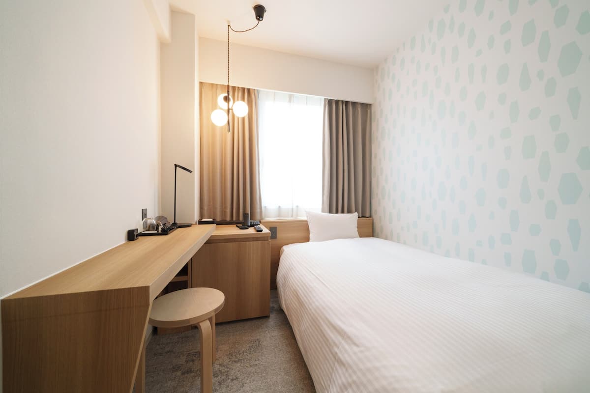A hotel room with a wooden desk, a small round table, a bed with white bedding, a nightstand, and a window with curtains.