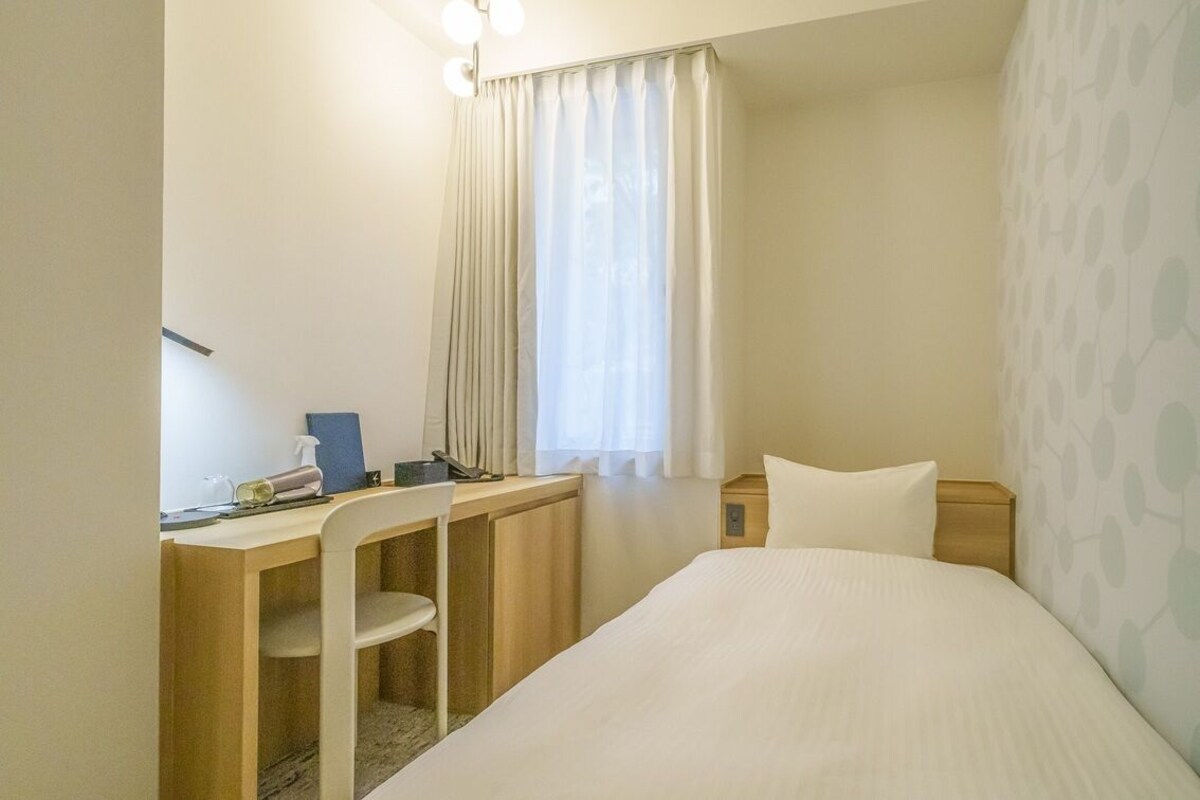 A hotel room with a bed, a desk, a chair, a nightstand, and a window with curtains.