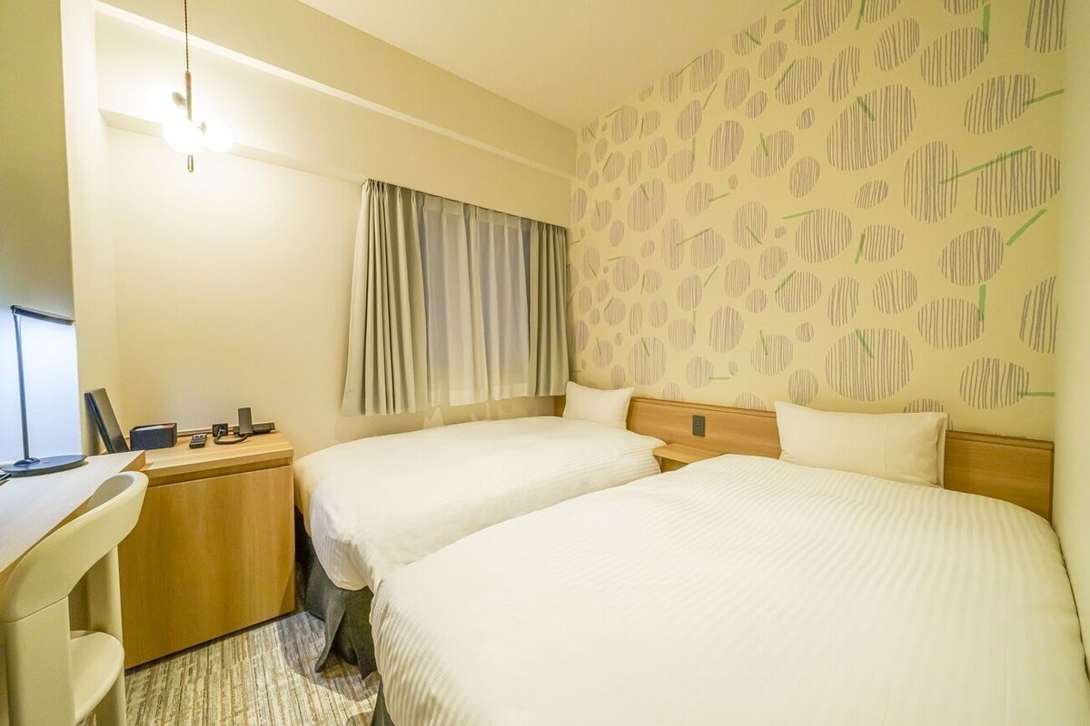 A hotel room with two beds, a desk, a chair, a lamp, and a window with curtains.