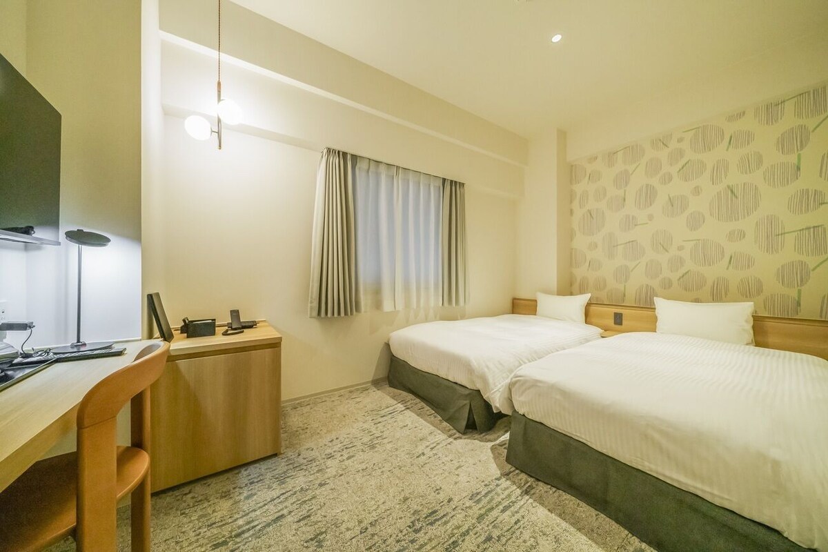 A hotel room with two beds, a desk, a chair, a lamp, and a window with curtains.