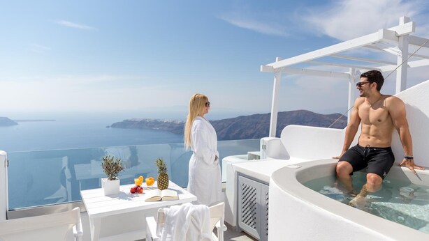 A couple on a balcony with a view of the sea, a hot tub, and a table with fruits and a book. - Unique Suites (Santorini)