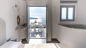 A bedroom with a bed, a bedside table, a nightstand, a window, and a view of the sea. - Unique Suites (Santorini)