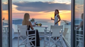 A balcony with two white chairs and a table, overlooking a sunset view. - Unique Suites (Santorini)