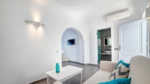 A modern living room with a white sofa, a small white table, and a teal vase. An arched doorway leads to a kitchen area. - Unique Suites (Santorini)