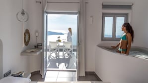 A room with a bed, a desk with a chair, a window, and a bathtub. - Unique Suites (Santorini)