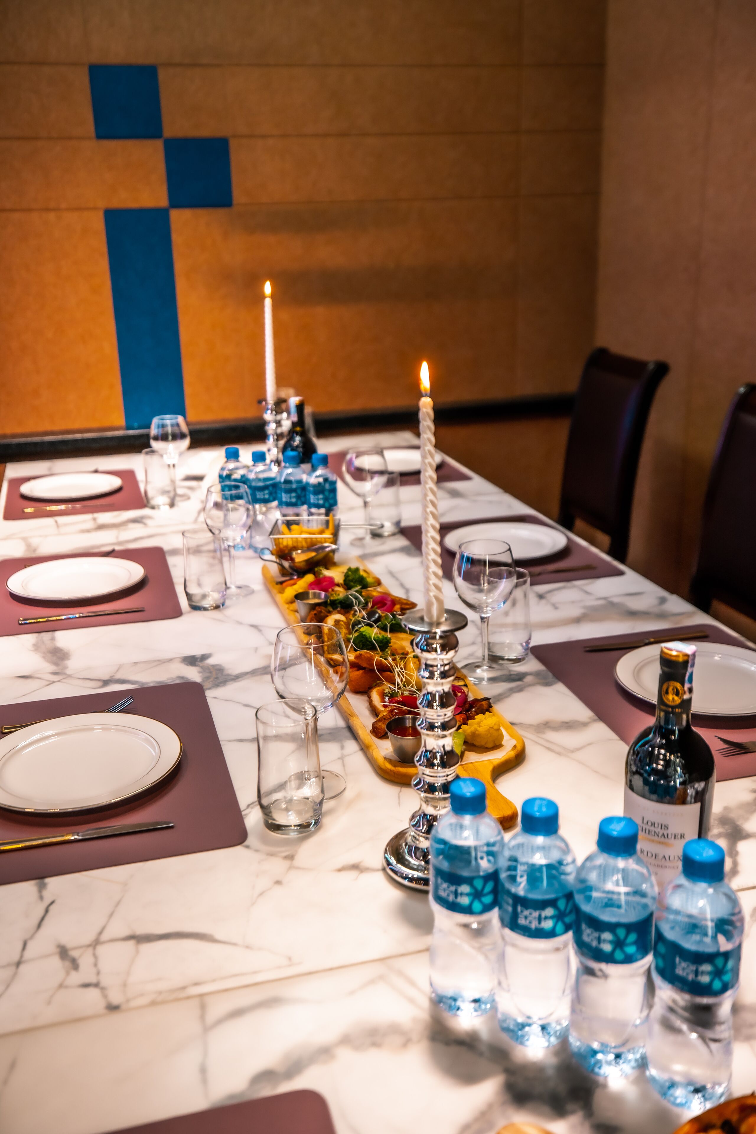 Una mesa de comedor dispuesta con platos, cubiertos, botellas de agua y un centro de mesa con velas.