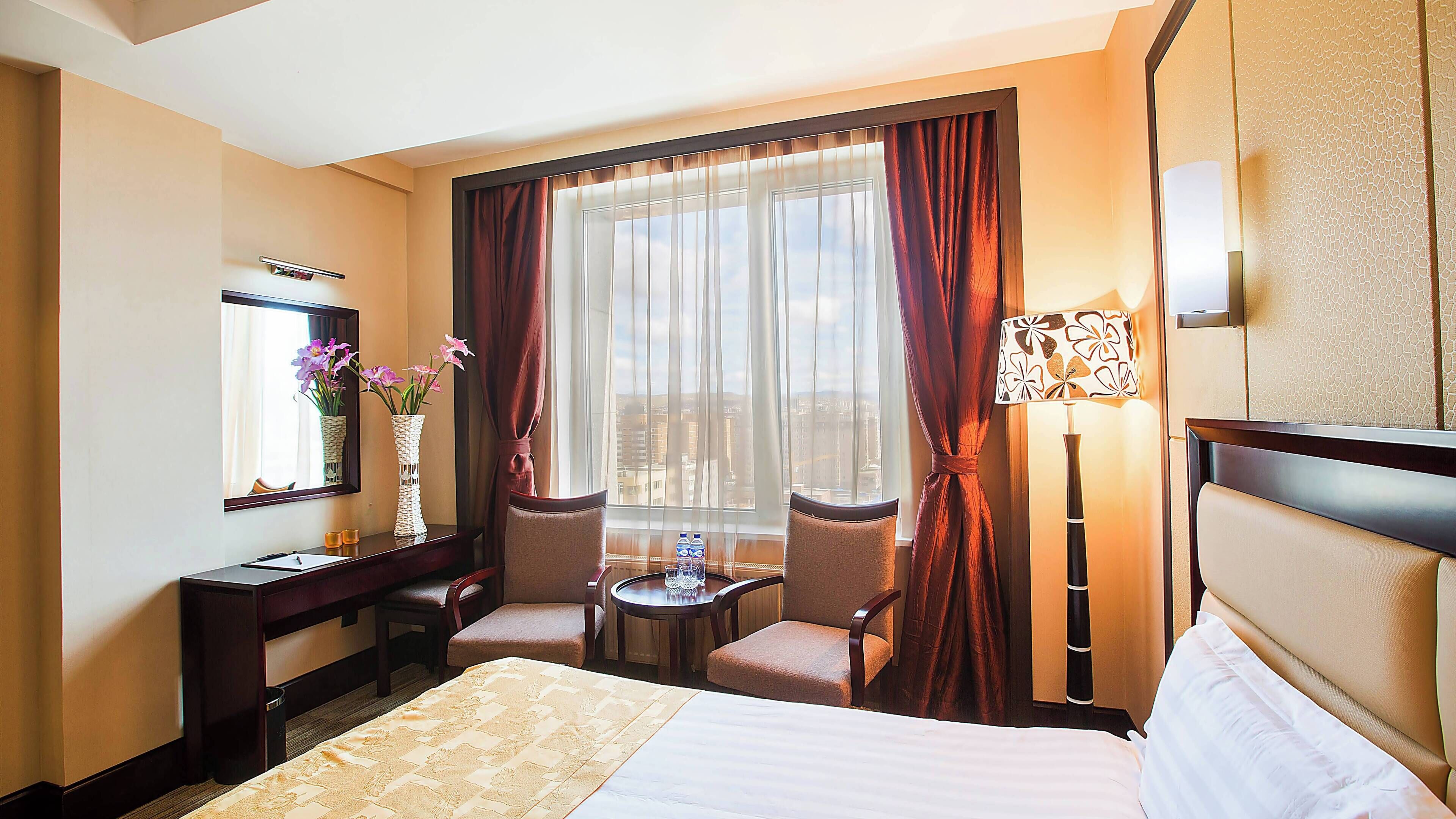 A hotel room with a bed, two chairs, a small table, a vase with flowers, a mirror, and a window with curtains.
