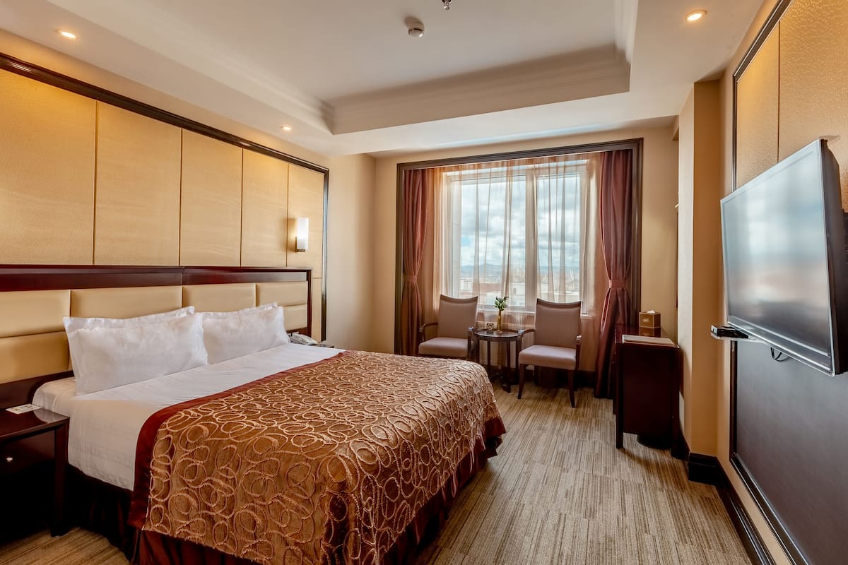 A hotel room with a large bed, a TV, a desk with two chairs, and a window with curtains.