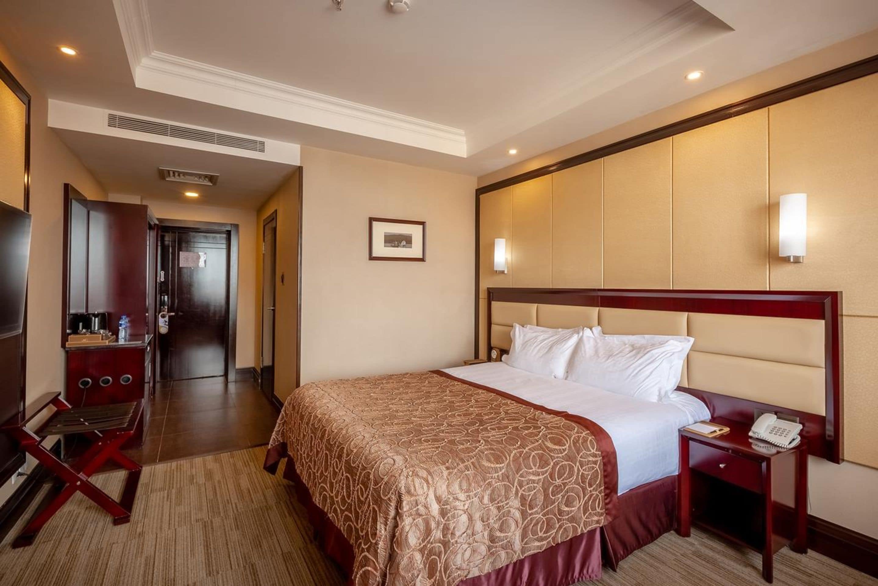 A hotel room with a large bed, a bedside table with a phone, a desk with a chair, a TV, and a door leading to a bathroom.
