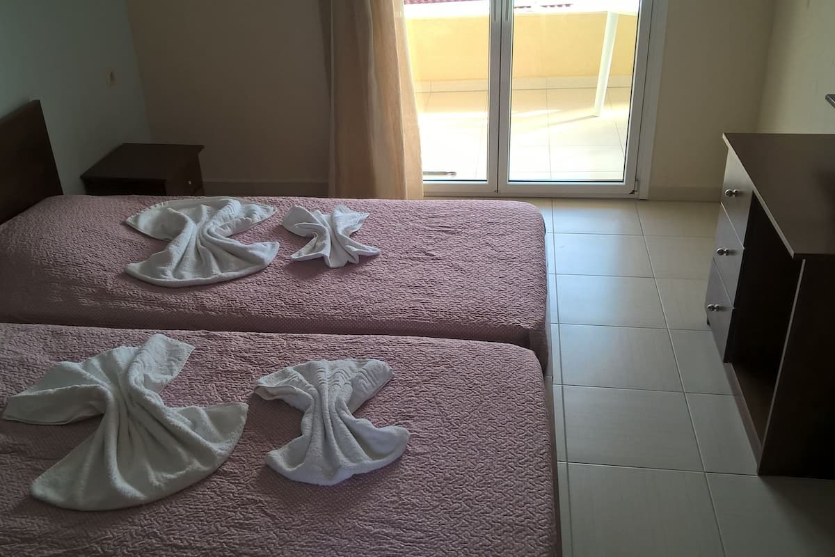 Habitación de hotel con dos camas, cada una con una colcha rosa texturizada y una toalla blanca dispuesta de forma decorativa. Hay una mesita de noche de madera y un armario a la derecha.