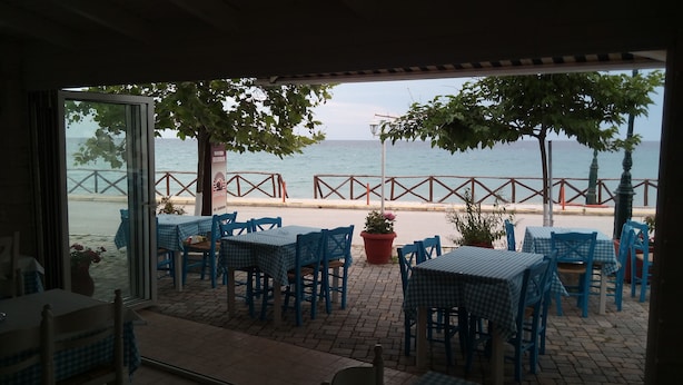 An outdoor seating area with checkered tables and chairs, a view of the sea, and a wooden fence along the walkway. - Kyparissis Rooms (Kassandra)