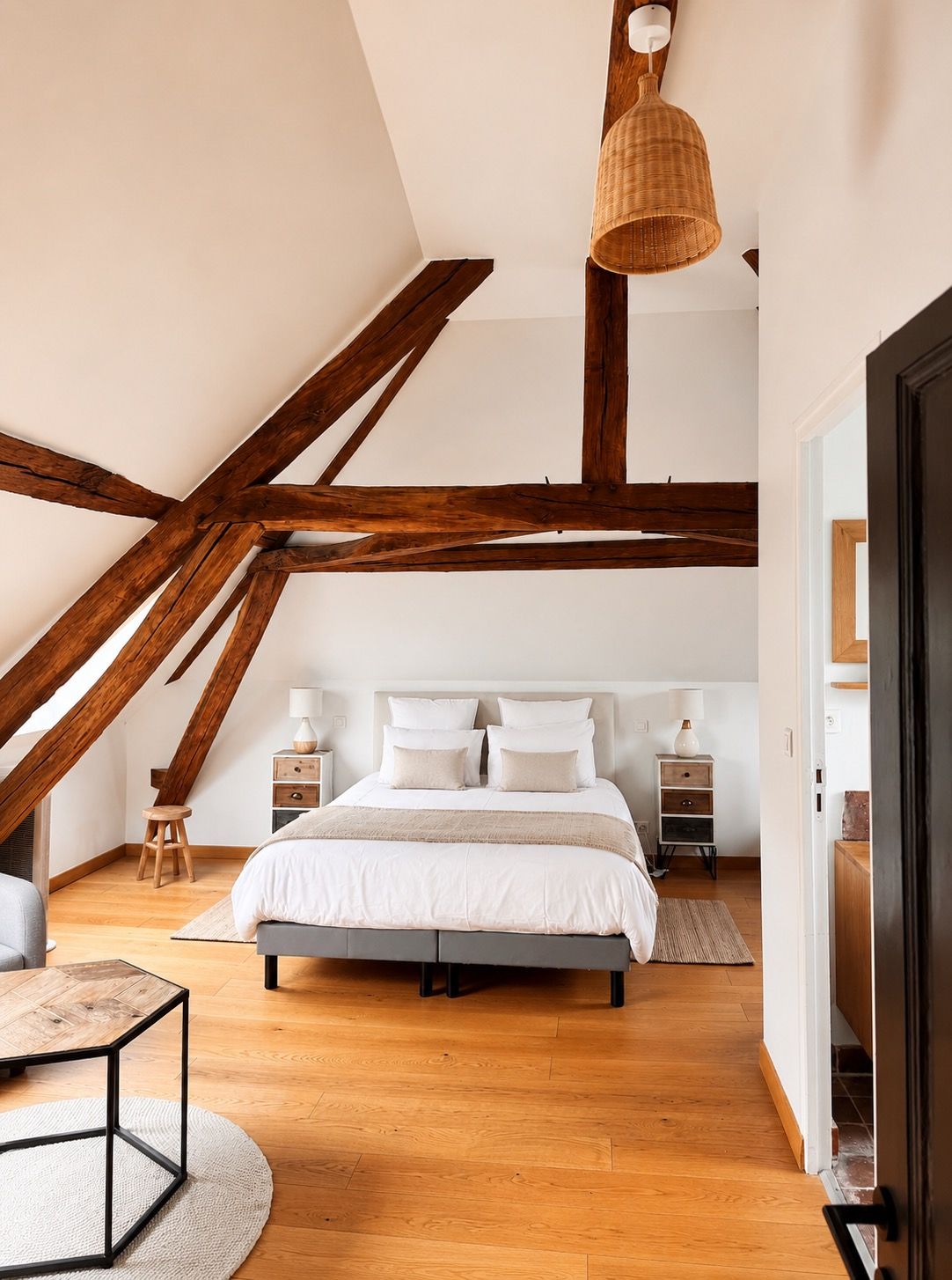 Un dormitorio con techo inclinado, vigas de madera a la vista, una cama con ropa blanca, mesitas de noche y una lámpara colgante.