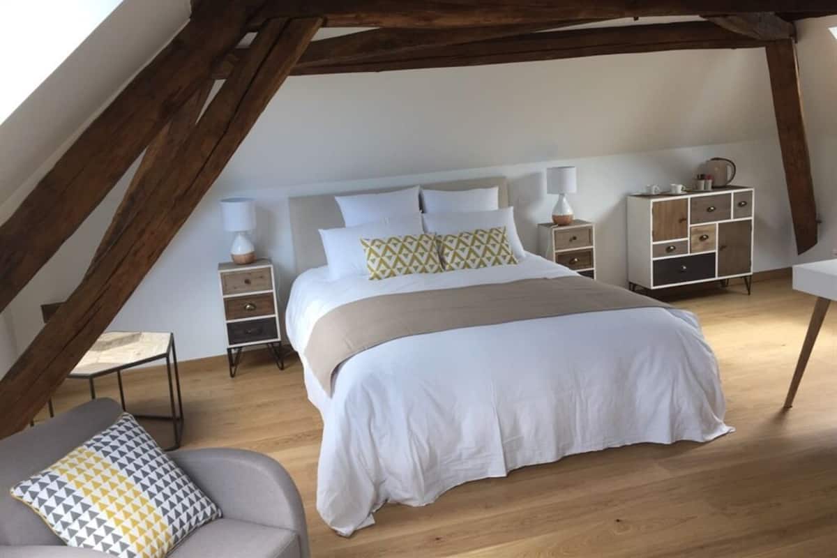 A bedroom with a large bed, wooden beams, a nightstand, a bedside table, a chair, and a small table.