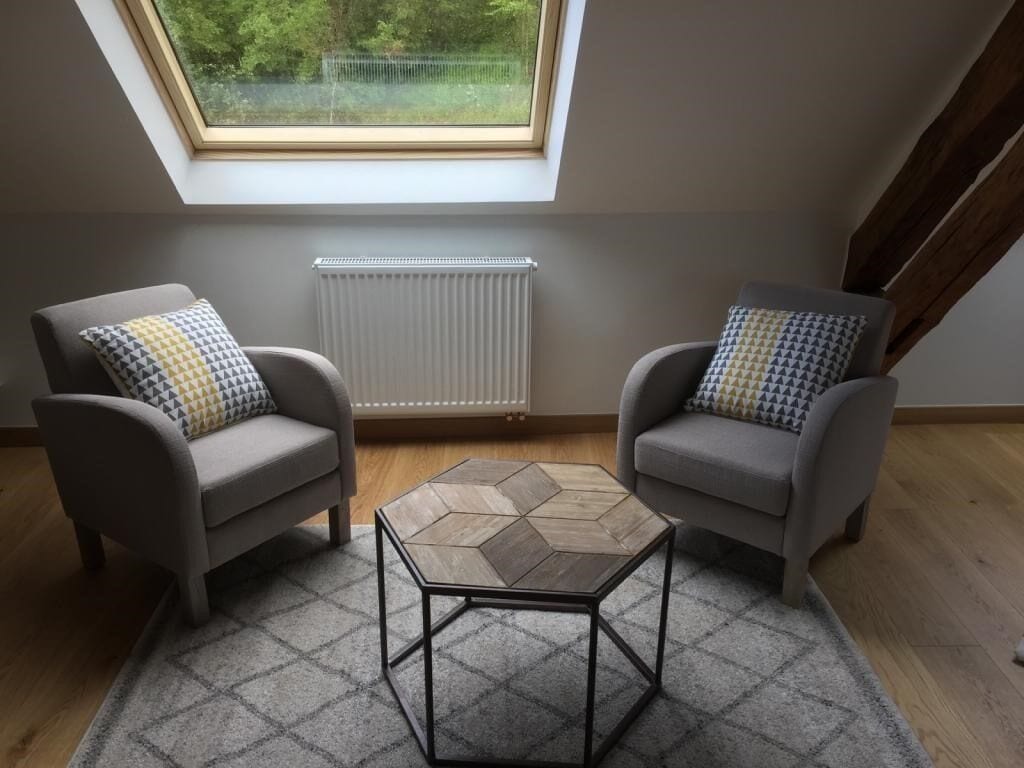 Una habitación con dos sillones, una pequeña mesa hexagonal, un radiador y un tragaluz.