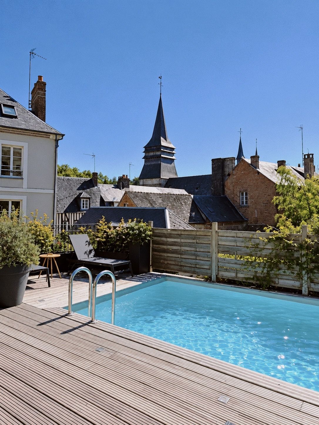En swimmingpool med ett trädäck, omgiven av hus och ett kyrktorn.