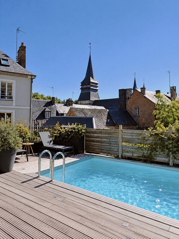A swimming pool with a wooden deck, surrounded by houses and a church spire.