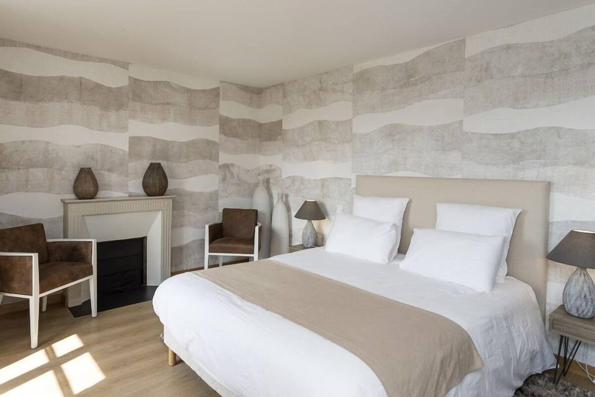 A modern bedroom with a large bed, a fireplace, two chairs, and a nightstand with a lamp.