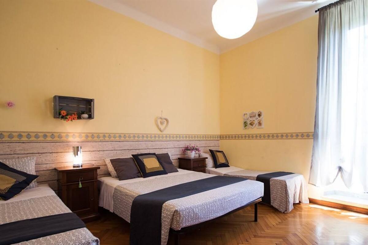 Een hotelkamer met drie eenpersoonsbedden, een houten vloer, een klein nachtkastje met een lamp en een raam met gordijnen.