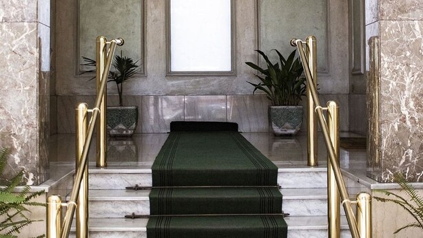 A marble staircase with green carpeted steps, flanked by gold railings, leading to a white door with a frosted glass panel.