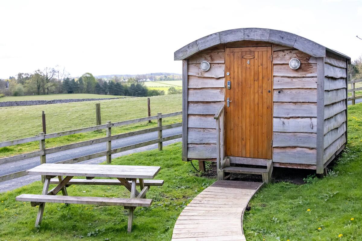 A rustic wooden outhouse with a wooden door and a number '4' on it, located next to a picnic table and a wooden pathway.