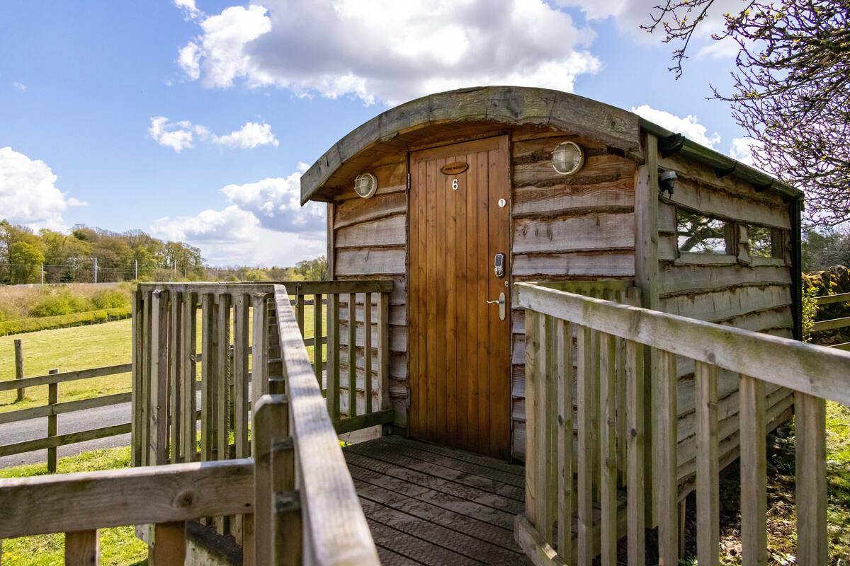 A wooden cabin with a wooden door and a number '6' on it, surrounded by a wooden fence and a wooden walkway leading to it.
